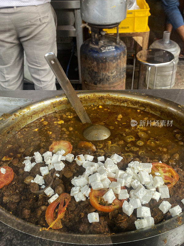 特写照片，一个认不出来的人正在准备街头小吃pindi chole(鹰嘴豆咖喱)，上面有奶酪块和西红柿片，放在大锅里，在市场摊位上的传统印度美味食物，高视图，重点在前景